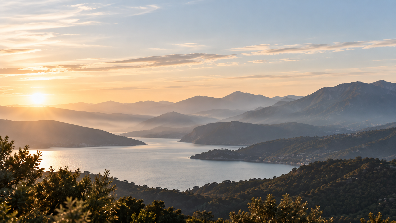 Lago entre montanhas em camadas, com vegetação próxima e horizontes distantes sob a luz suave do sol.